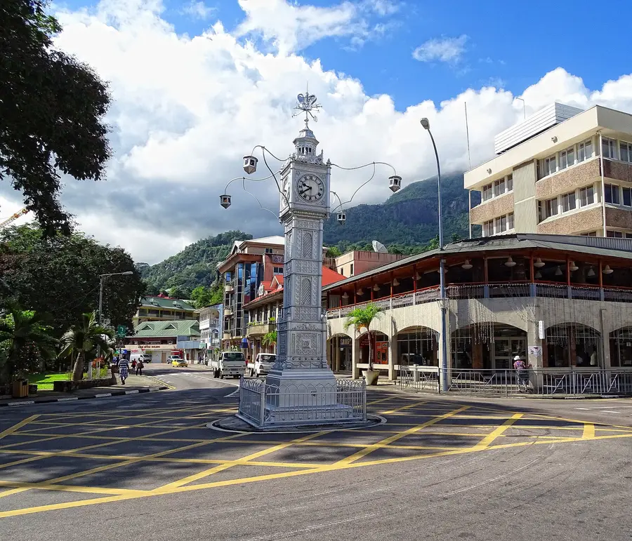 The Victoria Clock Tower in Victoria, Mahé, Seychelles, a silver clock column at a street junction with mountains behind it.