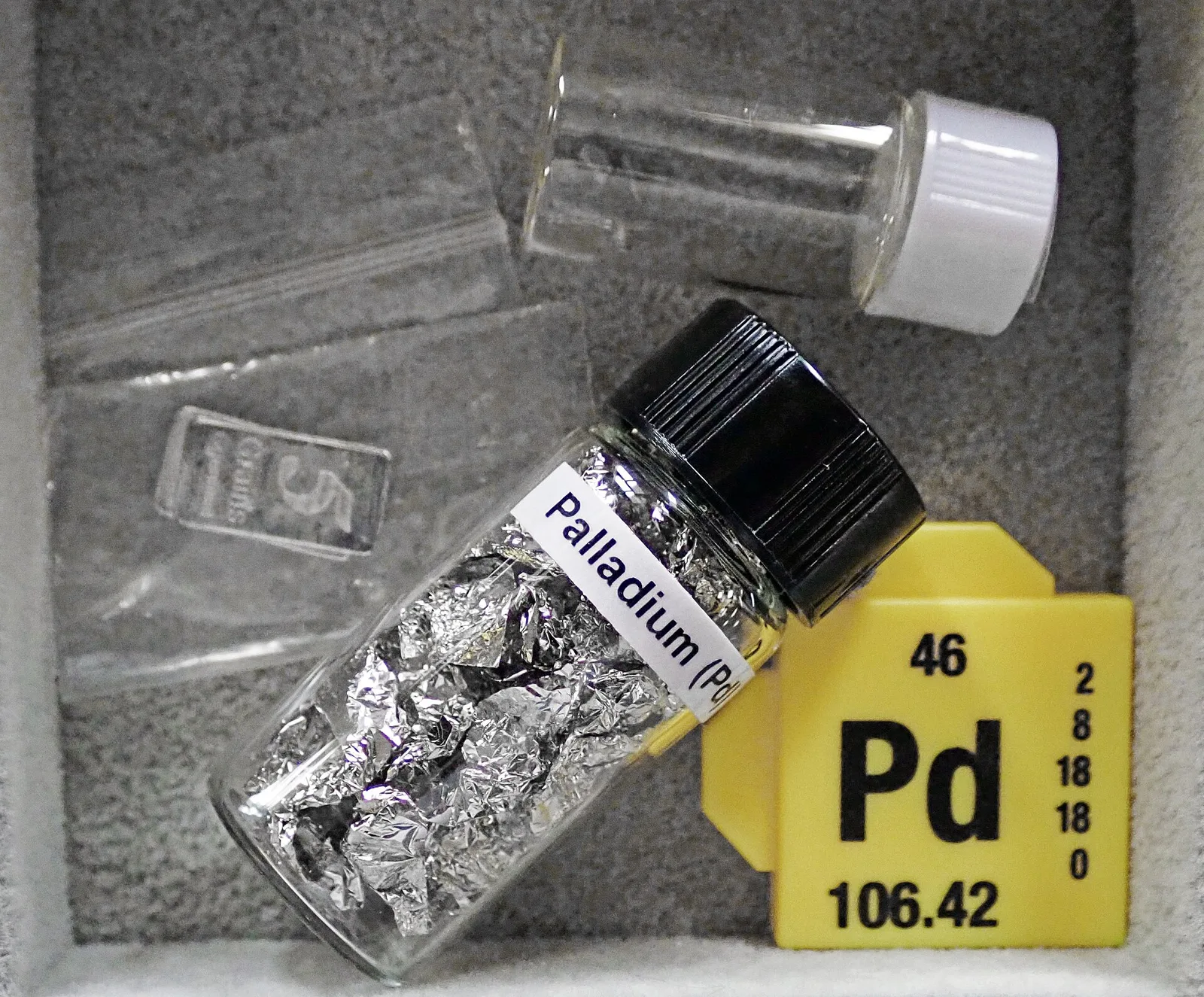 A labelled glass vial of palladium metal foil next to a periodic table tile for element 46, Pd, atomic mass 106.42