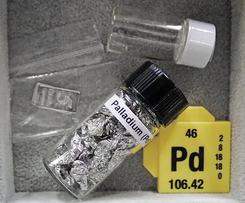 A labelled glass vial of palladium metal foil next to a periodic table tile for element 46, Pd, atomic mass 106.42