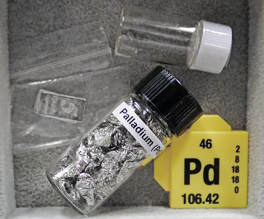 A labelled glass vial of palladium metal foil next to a periodic table tile for element 46, Pd, atomic mass 106.42