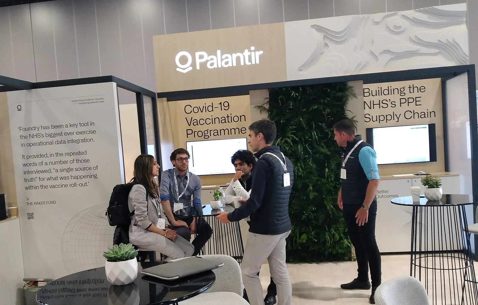 Staff and visitors talking at the Palantir Technologies exhibition stand, with the Palantir wordmark on the fascia and panels describing NHS data projects.