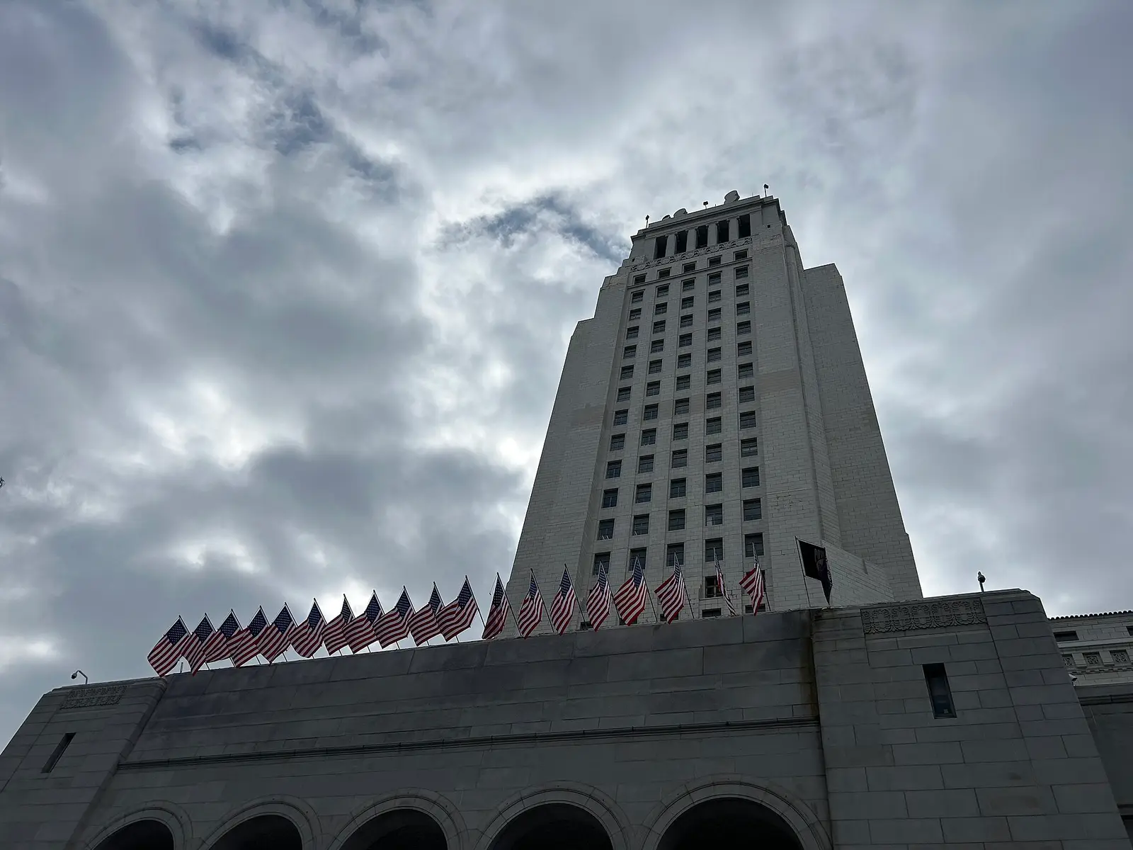Los Angeles City Hall tower with a row of American flags along its roofline under a cloudy sky