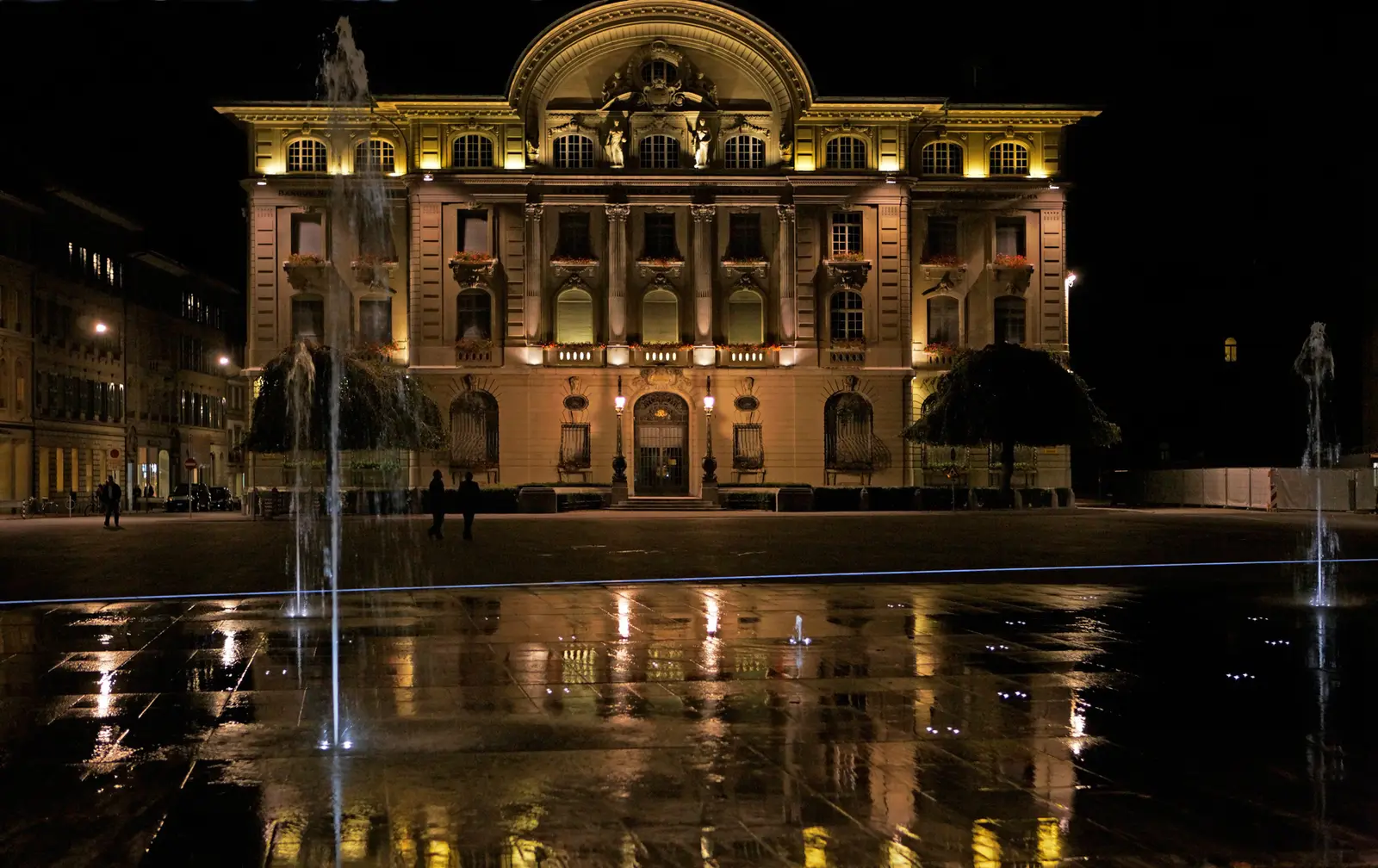 The Swiss National Bank building on Bundesplatz in Bern lit up at night, with the square water jets and wet paving in the foreground
