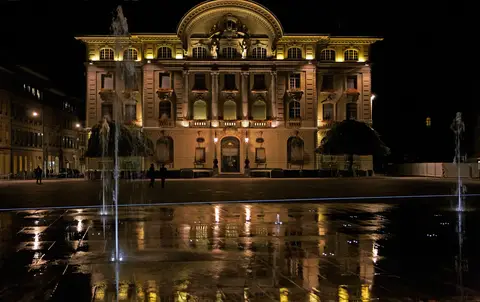 The Swiss National Bank building on Bundesplatz in Bern lit up at night, with the square water jets and wet paving in the foreground