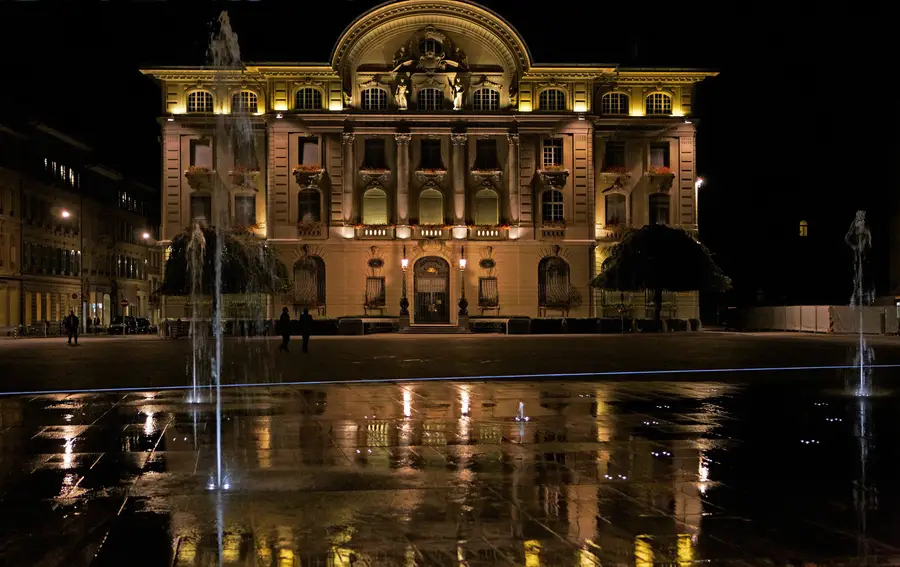 The Swiss National Bank building on Bundesplatz in Bern lit up at night, with the square water jets and wet paving in the foreground