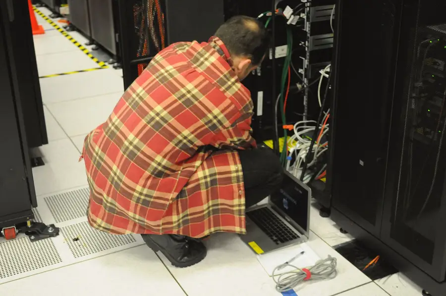 A technician crouches with a laptop at an open server rack in the NERSC data centre, working through cabling and network ports