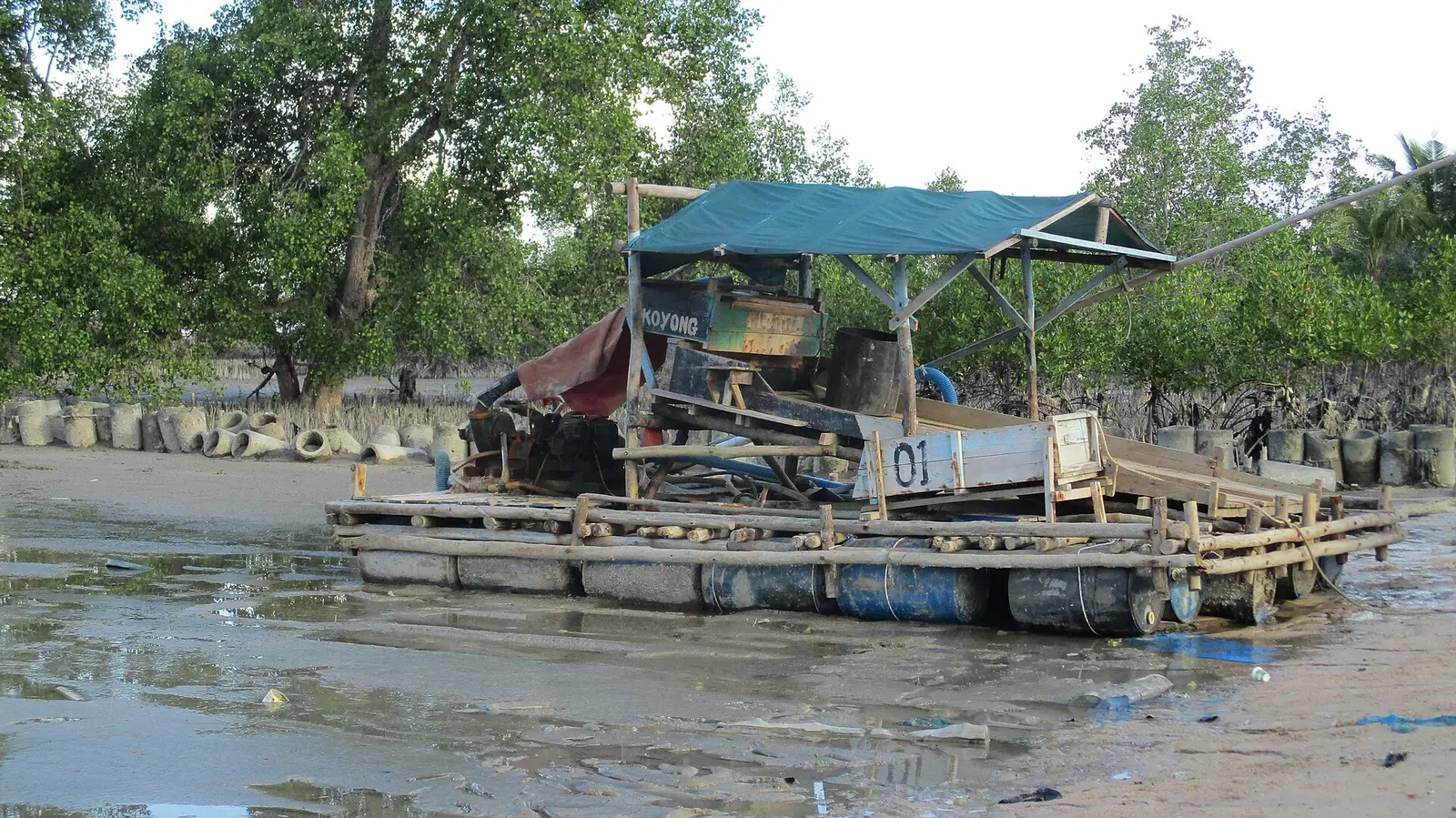 A floating tin mining pontoon moored on a tidal mudflat in Bangka Indonesia with a sluice box and pump mounted on a timber raft over oil drums